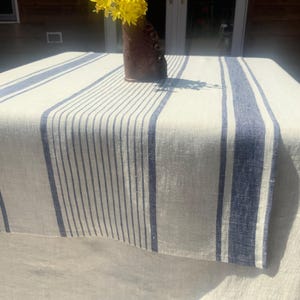 May include: A rectangular tablecloth with a cream-colored background and vertical blue stripes. A vase with yellow flowers sits on the table. The tablecloth is made of a linen-like material.