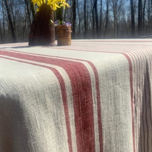 May include: A rectangular tablecloth featuring a woven off-white base with prominent vertical stripes in a deep red hue. The fabric displays a visible, textured weave pattern throughout, draping over the edges of a surface.