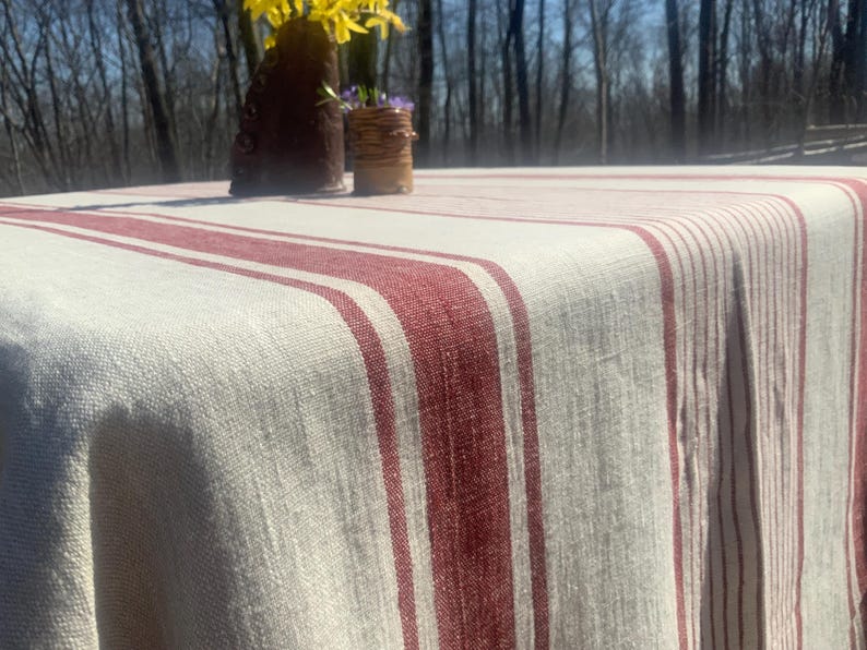 May include: A rectangular tablecloth featuring a woven off-white base with prominent vertical stripes in a deep red hue. The fabric displays a visible, textured weave pattern throughout, draping over the edges of a surface.
