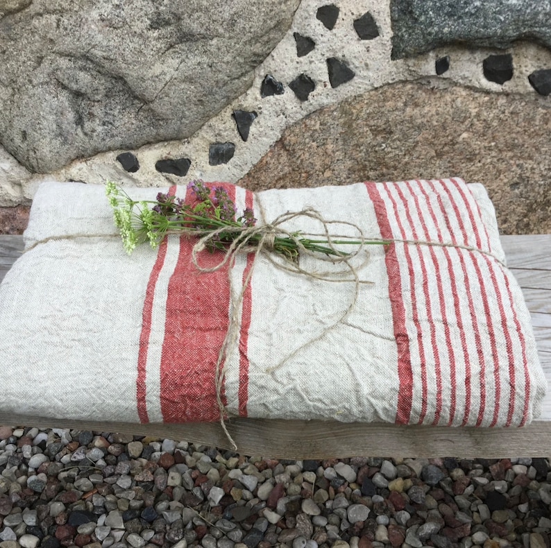 May include: A folded beige linen textile featuring prominent red vertical stripes of varying widths. The fabric is tied with a piece of natural twine and decorated with a small bundle of dried green and purple wildflowers resting on the surface.