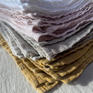 Puede incluir: Una pila de pa&ntilde;os cuadrados doblados con un patr&oacute;n de tejido de gofre texturizado. Los pa&ntilde;os est&aacute;n dispuestos en un degradado de colores, comenzando con blanco en la parte superior, seguido de rosa claro, gris suave y amarillo mostaza.