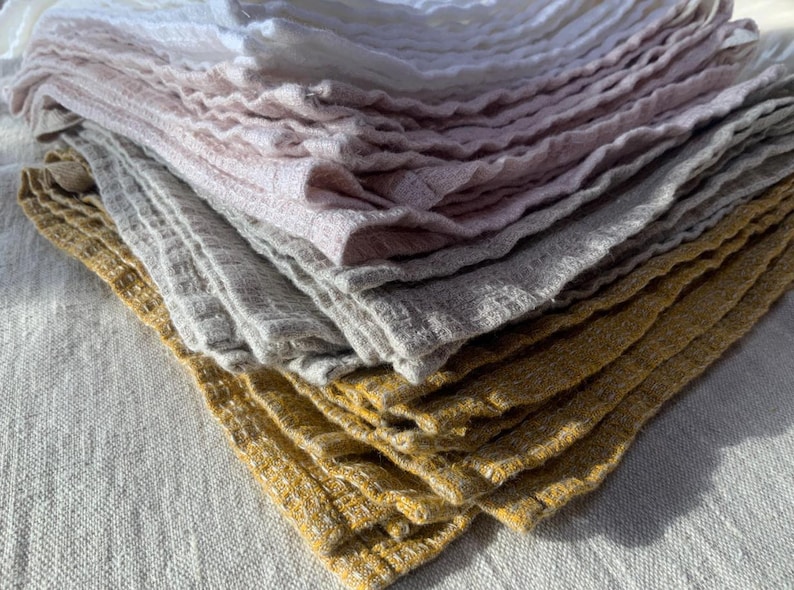 Puede incluir: Una pila de pa&ntilde;os cuadrados doblados con un patr&oacute;n de tejido de gofre texturizado. Los pa&ntilde;os est&aacute;n dispuestos en un degradado de colores, comenzando con blanco en la parte superior, seguido de rosa claro, gris suave y amarillo mostaza.