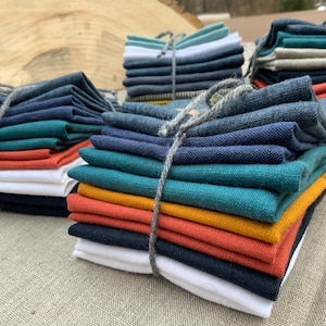 May include: Stacks of colorful fabric bundles tied with gray twine. The bundles feature a variety of colors, including white, teal, navy, orange, and yellow. The fabric appears to be linen or a similar material.