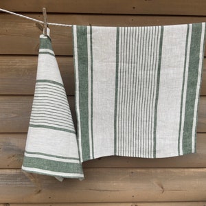 May include: Two rectangular kitchen towels made of off-white fabric with dark green stripes. One towel hangs folded, displaying horizontal stripes of varying widths, while the other hangs flat, featuring vertical stripes of different thicknesses.