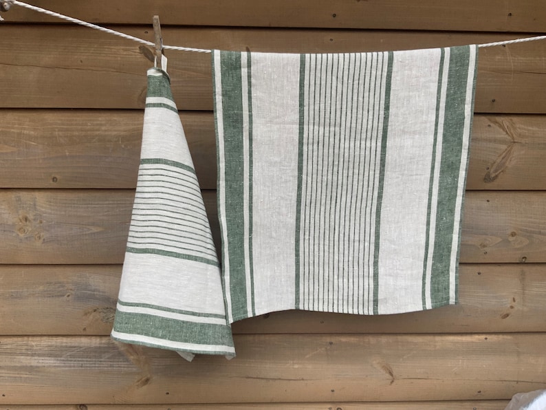 May include: Two rectangular kitchen towels made of off-white fabric with dark green stripes. One towel hangs folded, displaying horizontal stripes of varying widths, while the other hangs flat, featuring vertical stripes of different thicknesses.