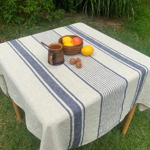 May include: A square beige tablecloth featuring thick and thin navy blue vertical stripes. On the fabric sits a dark brown textured ceramic mug with a wooden spoon, a wooden bowl containing two lemons and a peach, and three small brown walnuts.