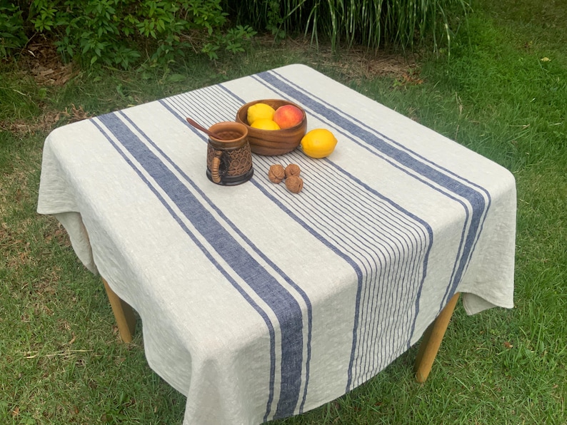 May include: A square beige tablecloth featuring thick and thin navy blue vertical stripes. On the fabric sits a dark brown textured ceramic mug with a wooden spoon, a wooden bowl containing two lemons and a peach, and three small brown walnuts.