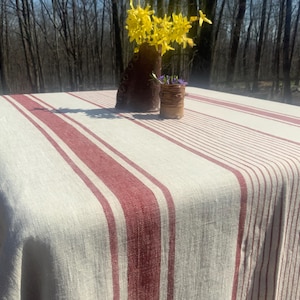 May include: A rectangular tablecloth featuring a natural beige base with prominent vertical stripes in a deep red hue. The fabric displays a textured weave, draping over the edges of a square table, with a dark brown vase holding yellow flowers.