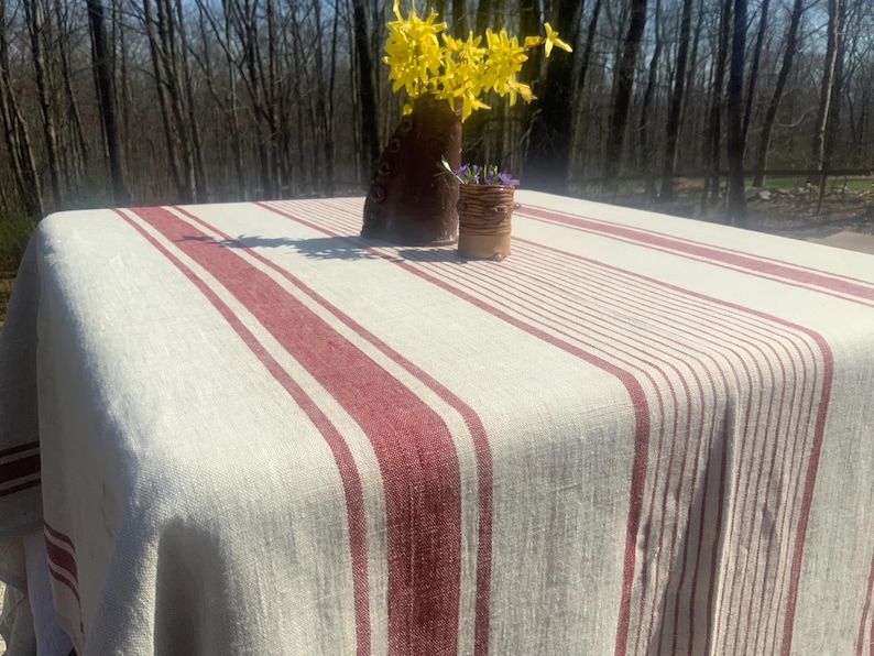May include: A rectangular tablecloth featuring a natural beige base with prominent vertical stripes in a deep red hue. The fabric displays a textured weave, draping over the edges of a square table, with a dark brown vase holding yellow flowers.