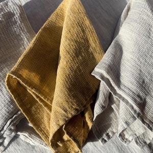 Puede incluir: Tres pa&ntilde;os de cocina plegados con una textura de tejido de gofre distintiva. El conjunto incluye un pa&ntilde;o de color amarillo mostaza intenso y dos pa&ntilde;os en un tono gris claro, todos mostrados con pliegues suaves y patrones de rejilla visibles.