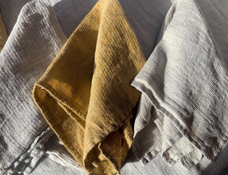 Puede incluir: Tres pa&ntilde;os de cocina plegados con una textura de tejido de gofre distintiva. El conjunto incluye un pa&ntilde;o de color amarillo mostaza intenso y dos pa&ntilde;os en un tono gris claro, todos mostrados con pliegues suaves y patrones de rejilla visibles.