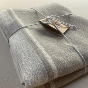 May include: A folded rectangular textile featuring a light beige and grey colour palette with distinct white and grey striped borders. The fabric is secured with a thin white string tied in a bow around a small, rectangular brown paper tag.