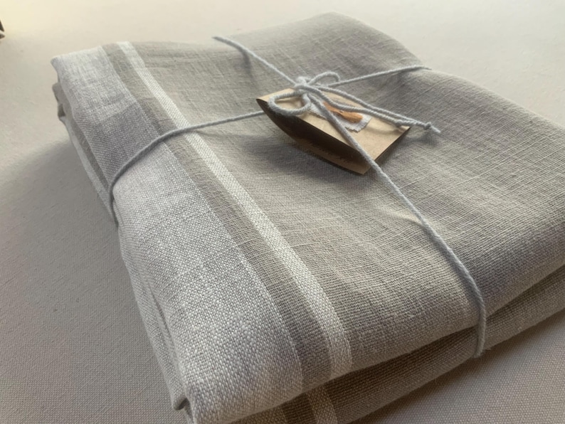 May include: A folded rectangular textile featuring a light beige and grey colour palette with distinct white and grey striped borders. The fabric is secured with a thin white string tied in a bow around a small, rectangular brown paper tag.