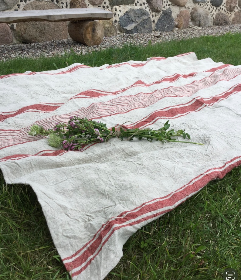 May include: A rectangular off-white fabric cloth featuring horizontal red stripes of varying widths. The textile has a visible wrinkled texture and is draped across the ground. A small bundle of green stems with purple and white flowers is tied with twine.