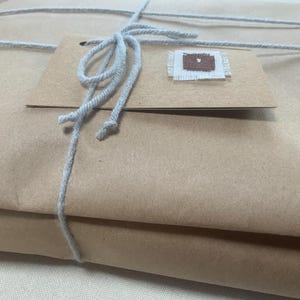 May include: A rectangular package wrapped in plain brown paper, secured with light grey string tied in a bow. A rectangular brown paper tag is attached to the string, featuring a small, square, brown and white fabric patch sewn onto it.