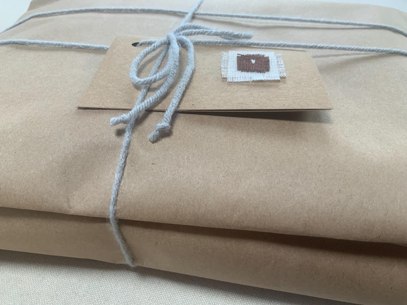 May include: A rectangular package wrapped in plain brown paper, secured with light grey string tied in a bow. A rectangular brown paper tag is attached to the string, featuring a small, square, brown and white fabric patch sewn onto it.