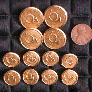 May include: A collection of ten gold-toned metal buttons with a stylized horn design. The buttons are arranged in a circular pattern on a black fabric background.