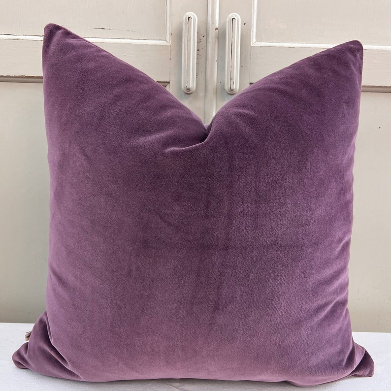 May include: A plush purple velvet cushion with a square shape and a simple design.