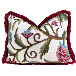 May include: A rectangular throw pillow with a white base fabric featuring colorful floral embroidery in shades of pink, blue, and green. The edges of the pillow are finished with a thick, textured deep red fringe trim.