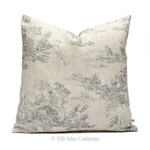 May include: A beige linen pillow with a blue and white toile pattern featuring a pastoral scene with people, trees, and animals. The pillow has a subtle texture and a slightly distressed look.
