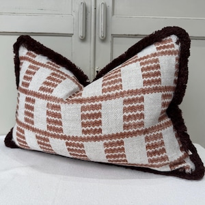 May include: Rectangular cushion with a white and brown plaid design. It features a dark brown fringe trim around the edges. The cushion is placed on a white surface.