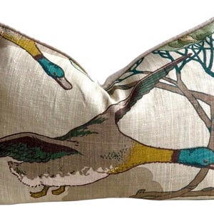 May include: A rectangular throw pillow with a beige linen-like texture featuring a printed pattern of mallard ducks in flight. The ducks have green heads, yellow necks, and brown wings. The pillow is finished with a light brown piped edge.