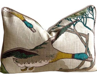 May include: A rectangular throw pillow with a beige linen-like texture featuring a printed pattern of mallard ducks in flight. The ducks have green heads, yellow necks, and brown wings. The pillow is finished with a light brown piped edge.