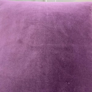 May include: A close-up of a soft, plush purple cushion. The fabric has a subtle texture and a slightly faded appearance.