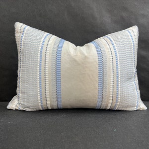 May include: Rectangular decorative pillow with a light gray center panel and vertical stripes in shades of blue, white, and gray. The pillow has a textured, woven appearance and is designed for home decor.