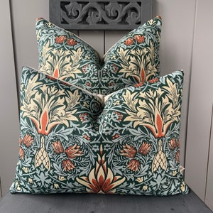 May include: Two decorative throw cushions with a floral pattern in shades of green, orange, and cream. The cushions are made of a soft, textured fabric and feature a repeating design of stylised flowers and leaves.
