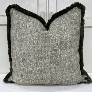 Puede incluir: Un cojín decorativo cuadrado con un patrón texturizado gris y blanco. El cojín tiene un borde con flecos verde oscuro. El color neutro del cojín complementaría muchos estilos de decoración del hogar. Aproximadamente 40 cm x 40 cm.