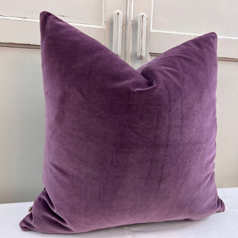 May include: A plush purple velvet cushion with a square shape. The cushion is soft and has a smooth texture.