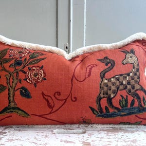 May include: A rectangular throw cushion in a burnt orange shade featuring a printed design of a stylised tree with red flowers and a chequered deer. The cushion is finished with a thick, cream-coloured fringe trim along all four edges of the fabric.