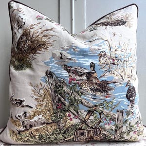 May include: Cushion with a nature-themed design. The print depicts ducks in a pond, with reeds, rocks, and a wooden fence. The cushion has a cream background and a dark brown trim. The design is reminiscent of a vintage toile.