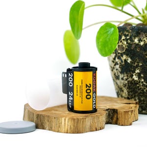 May include: A roll of Kodak Gold 200 color negative film with a yellow label and black text. The film canister is positioned next to its translucent white plastic container and a gray cap, featuring 24 exposures and 35mm film format specifications.