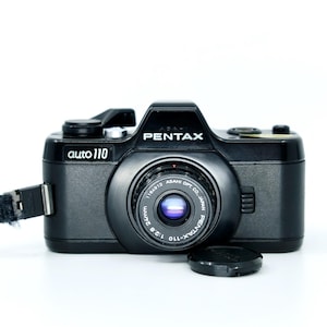 May include: A black Pentax auto 110 film camera with a 24mm 1:2.8 lens, featuring a black wrist strap attached to the side and a small circular lens cap resting in front of the camera body on a plain white background.