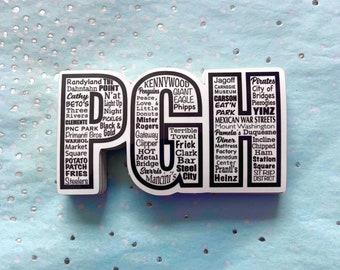 May include: A white and black sticker in the shape of the letters "PGH" with a list of Pittsburgh landmarks and attractions. The sticker includes text such as "Kennywood," "Pirates," and "Strip District."