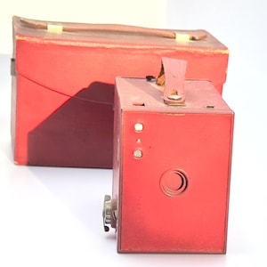 May include: A red vintage box camera with a leather carrying case. The camera has a round lens and a leather strap.