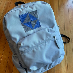 May include: A light gray backpack with a front pocket and a blue and gray patchwork design on the front flap.