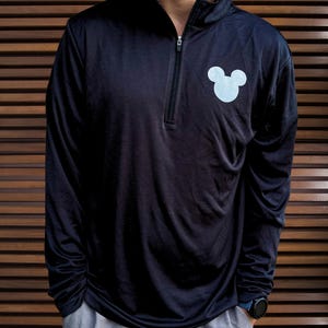 May include: Navy blue long-sleeve pullover shirt with a quarter-zip collar and a light blue Mickey Mouse head graphic on the chest. The shirt is paired with gray shorts. The person is wearing a black watch.