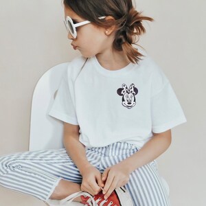 May include: A white t-shirt with a Minnie Mouse graphic, paired with blue and white striped pants and red sneakers. The child is wearing white sunglasses and sitting on a white chair.