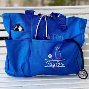 Puede incluir: Una bolsa de lona azul con un estetoscopio y el nombre "Taylor" bordado. La bolsa tiene varios bolsillos y un cierre de cremallera. La bolsa est&aacute; sobre una superficie de madera blanca.