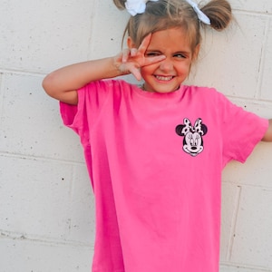 May include: A bright pink t-shirt featuring a Minnie Mouse graphic on the front. The graphic is a black and white image of Minnie Mouse's face with a polka dot bow. The shirt is worn by a young person.