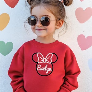 May include: A red sweatshirt featuring a black and white Mickey Mouse head design with the name "Evelyn" in white. The sweatshirt is being worn by a young girl with brown hair and sunglasses.