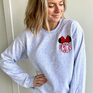 Puede incluir: Una sudadera gris con orejas de Mickey Mouse y un lazo bordados en rojo y negro en el pecho izquierdo. La sudadera tiene cuello redondo y mangas largas.