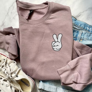 May include: A mauve-colored sweatshirt with a white and black peace sign graphic. The sweatshirt is paired with blue denim jeans and white canvas sneakers with red accents. The jeans have the text "Gap 1969" printed on the pocket.