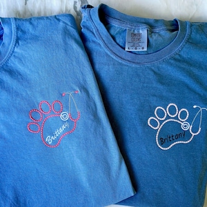 May include: Two blue Comfort Colors t-shirts. One shirt has a pink paw print design with a stethoscope and the name "Brittany" embroidered in pink. The other shirt has a white paw print design with a stethoscope and the name "Brittany" embroidered in white.