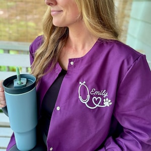 May include: A purple medical scrub top with a white embroidered stethoscope, heart, and paw print. The name "Emily" is embroidered on the top.