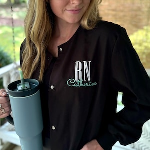 May include: A black scrub top with a white embroidered RN and the name Catherine in teal. The person is holding a teal travel mug with a straw.