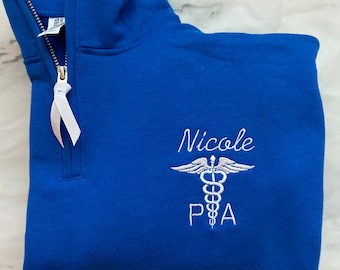 May include: A royal blue 1/4 zip sweatshirt with a white embroidered medical caduceus symbol and the text "Nicole PA".